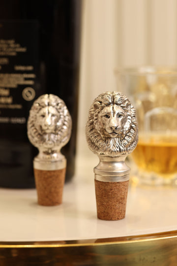 Two silver-plated brass lion head wine bottle stoppers with cork, beside a bottle and a glass