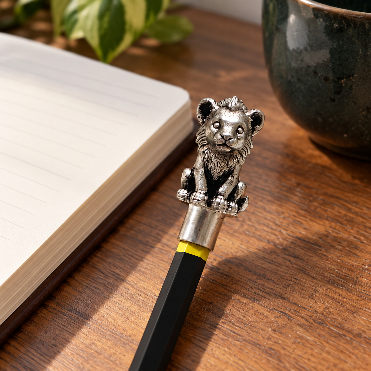 Sheru lion cub pencil topper fitted on a pencil beside an open notebook on a wooden desk